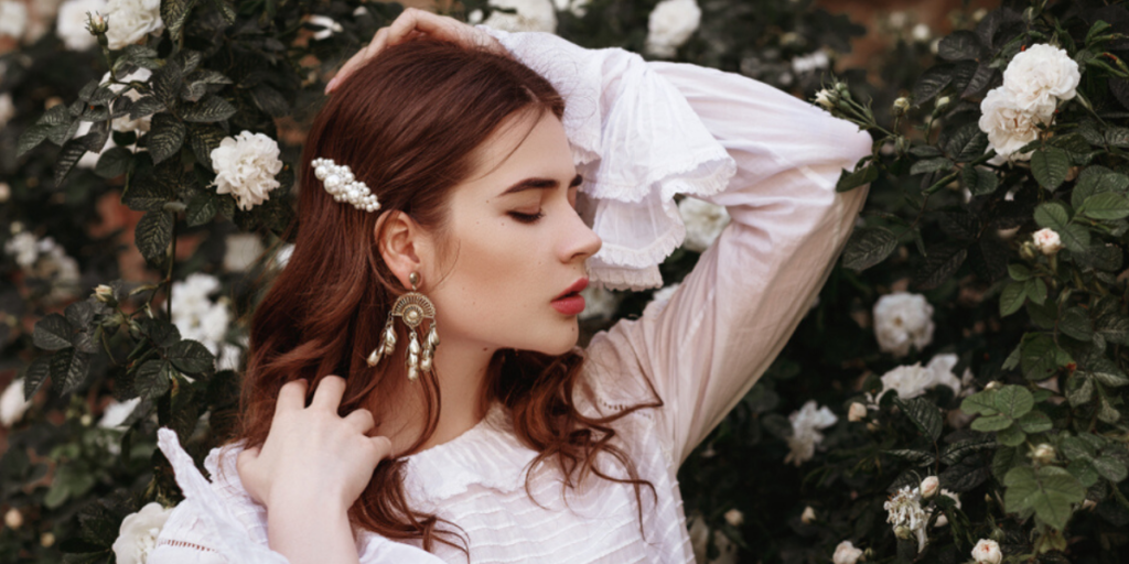 Brunette girl in a whit shirt and pearl barrette stands in front of white flowers.